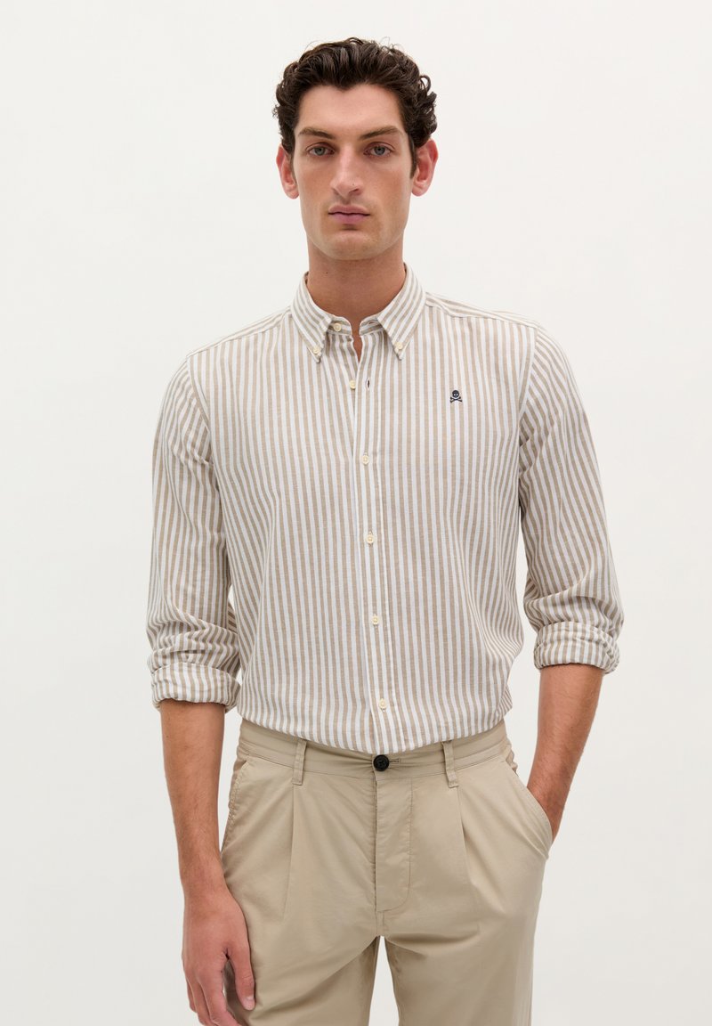 Camicia button-down con strisce verticali beige e bianche, maniche lunghe arrotolate, e un piccolo logo sul petto. Abbinata a pantaloni beige.