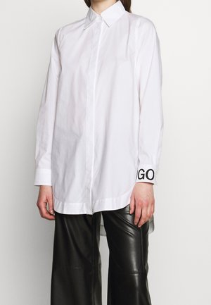 Personne portant une chemise blanche à manches longues boutonnée avec le texte "GO" sur le poignet et un pantalon en cuir noir sur fond uni.