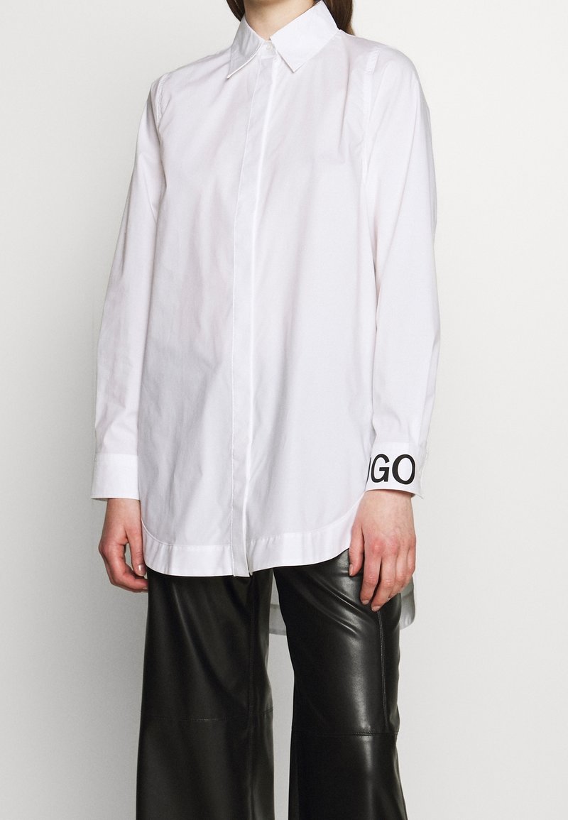 Personne portant une chemise blanche à manches longues boutonnée avec le texte "GO" sur le poignet et un pantalon en cuir noir sur fond uni.