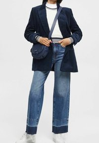 Marineblaues Cord-Blazer über einem weißen Strickpullover, kombiniert mit weit geschnittenen blauen Jeans und weißen Sneakern. Eine dunkelblaue Umhängetasche rundet den Look ab.