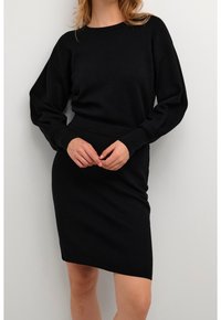 Femme portant une robe noire ajustée à manches longues avec une taille côtelée et des manches légèrement bouffantes, se tenant contre un fond clair uni.