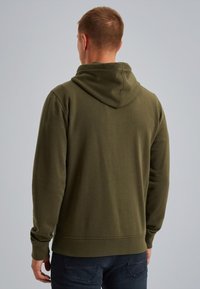 Uomo che indossa una felpa con cappuccio verde oliva a maniche lunghe, in piedi con la schiena rivolta verso la macchina fotografica su uno sfondo grigio uniforme.