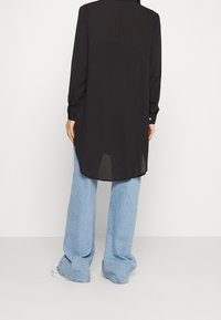 Persona che indossa una lunga camicia nera con bottoni e jeans a gamba larga blu chiaro, in piedi su uno sfondo chiaro e semplice.