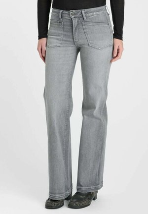 Persoon draagt lichtgrijze rechte jeans met voorzakken en zwarte schoenen met hak tegen een effen achtergrond.