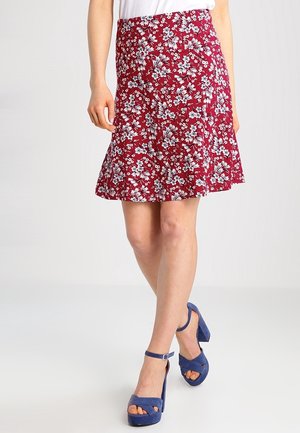 Mujer que lleva una falda roja con flores hasta la rodilla y sandalias azules de plataforma con tacón alto, de pie con las piernas cruzadas.