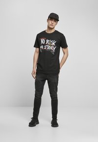 Camiseta de algodón negra con texto gráfico en blanco y rojo "NO RISK NO STORY". Combinada con jeans oscuros y zapatos negros; fondo liso.