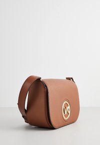 Borsa a tracolla in pelle marrone dalla forma arrotondata, con un logo circolare dorato sulla parte frontale e una tracolla regolabile. Texture liscia.