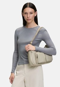 Borsa a spalla in pelle beige con molteplici tasche con zip; texture liscia; forma morbida e arrotondata; indossata con un top grigio a coste aderente.