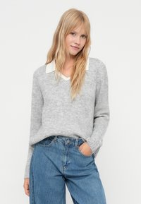 Blonde sieviete ar vaļīgiem matiem, uzvilkusi gaiši pelēku v-neck džemperi pār baltu krekla pusi un ziliem džinsiem, stāvot ar vienu roku kabatā.