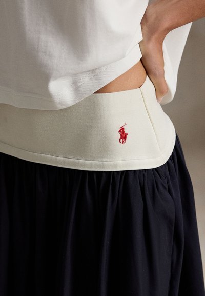 Λευκό βαμβακερό T-shirt με στρώση πάνω από ελαστική μπεζ ζώνη μέσης με κόκκινο κεντητό λογότυπο, σε συνδυασμό με μαύρη φουσκωτή φούστα με πιέτες.