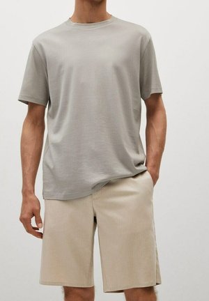 Mann trägt ein schlichtes hellgraues T-Shirt mit kurzen Ärmeln, leicht in beige knielange Shorts gesteckt, vor weißem Hintergrund.