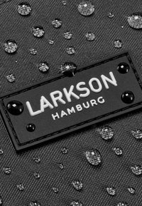 Schwarzer Stoff mit Wassertropfen, versehen mit einem rechteckigen Etikett, auf dem in erhabenen weißen Buchstaben und glänzenden schwarzen Akzenten "LARKSON HAMBURG" steht.