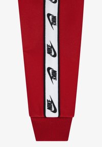 Pantalones deportivos rojos con una franja lateral blanca que presenta repetidos logotipos negros de "Nike" y puños acanalados. La tela es suave con un acabado mate.