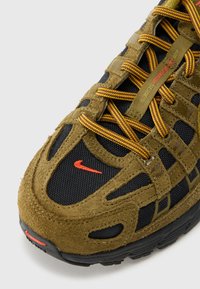 Buty sportowe Nike z brązową cholewką ze zamszu, czarnymi panelami z siateczki i pomarańczowymi akcentami. Teksturowane sznurowadła w kolorach żółtym i brązowym. Solidna czarna gumowa podeszwa.