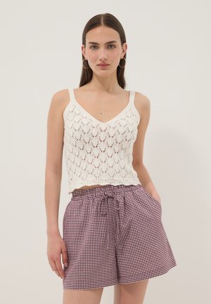 Joven mujer con top blanco de crochet y shorts de cintura alta a cuadros rojos y blancos, de pie con las manos en los bolsillos.