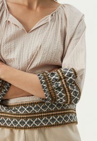 Vaaleanbeige, tekstuuriltaan mielenkiintoinen pusero, jossa on V-pääntie ja kolmivartiset hihat. Hihansuissa on hienostunutta mustaa ja oranssia kukkakirjontaa.