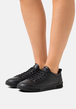 Jalat mengenakan sepatu sneaker hitam rendah dengan tali, terbuat dari tekstur kulit yang halus, berdiri di atas latar belakang putih.