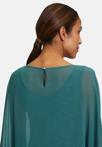 Haut en chiffon teal avec un col arrondi, présentant une découpe en forme de trou de serrure à l'arrière et une petite fermeture à bouton. Texture lisse et légère.
