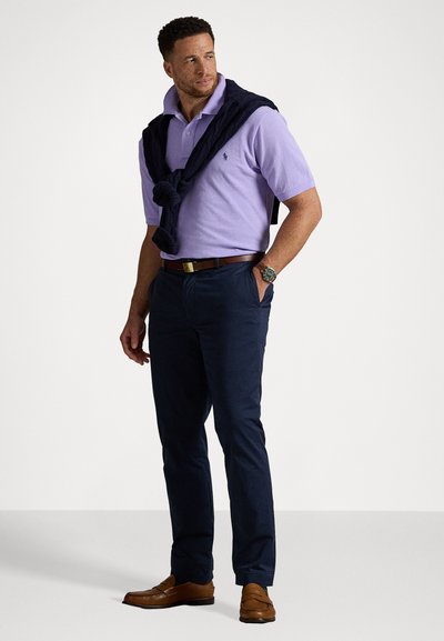 Gai violetā polo krekls ar tumši zilu džemperi, kas uzlikts uz pleciem, apvienots ar tumši ziliem biksēm un brūnām loafers.