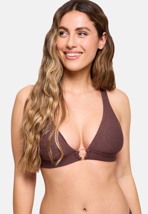 Mujer sonriente con cabello largo y ondulado, vistiendo un top de bikini marrón en forma de triángulo con un detalle de anillo frontal, mirando hacia un lado sobre un fondo liso.