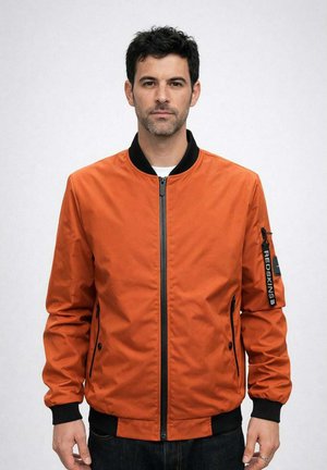 Homme portant une veste aviateur orange avec des poignets et une fermeture éclair noirs, debout devant un fond gris clair uni.