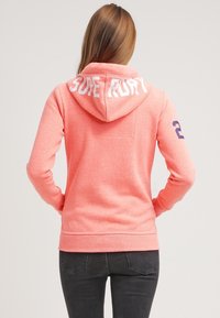 Donna che indossa un felpa corallo con la scritta "SURF RURY" sul cappuccio e il numero "2" blu navy sul maniche destra, girata di spalle.