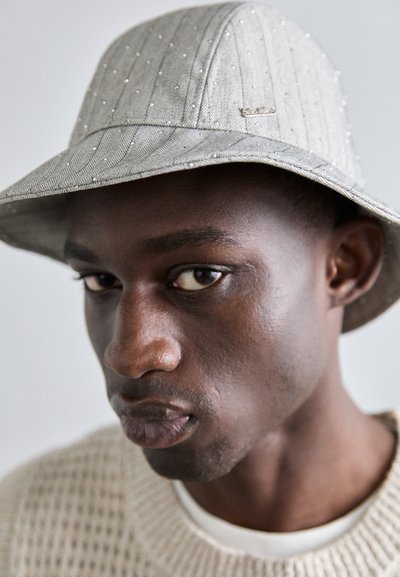 Chapeau seau gris avec de subtiles rayures, orné de petits accents brillants. Le bord est légèrement incliné vers le bas, associé à un pull en tricot texturé.