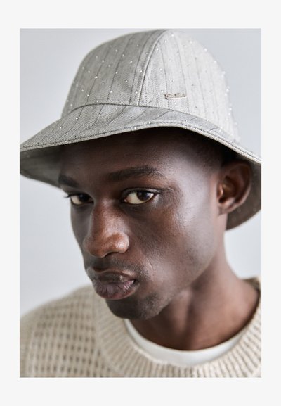 Chapeau seau gris avec de subtiles rayures, orné de petits accents brillants. Le bord est légèrement incliné vers le bas, associé à un pull en tricot texturé.