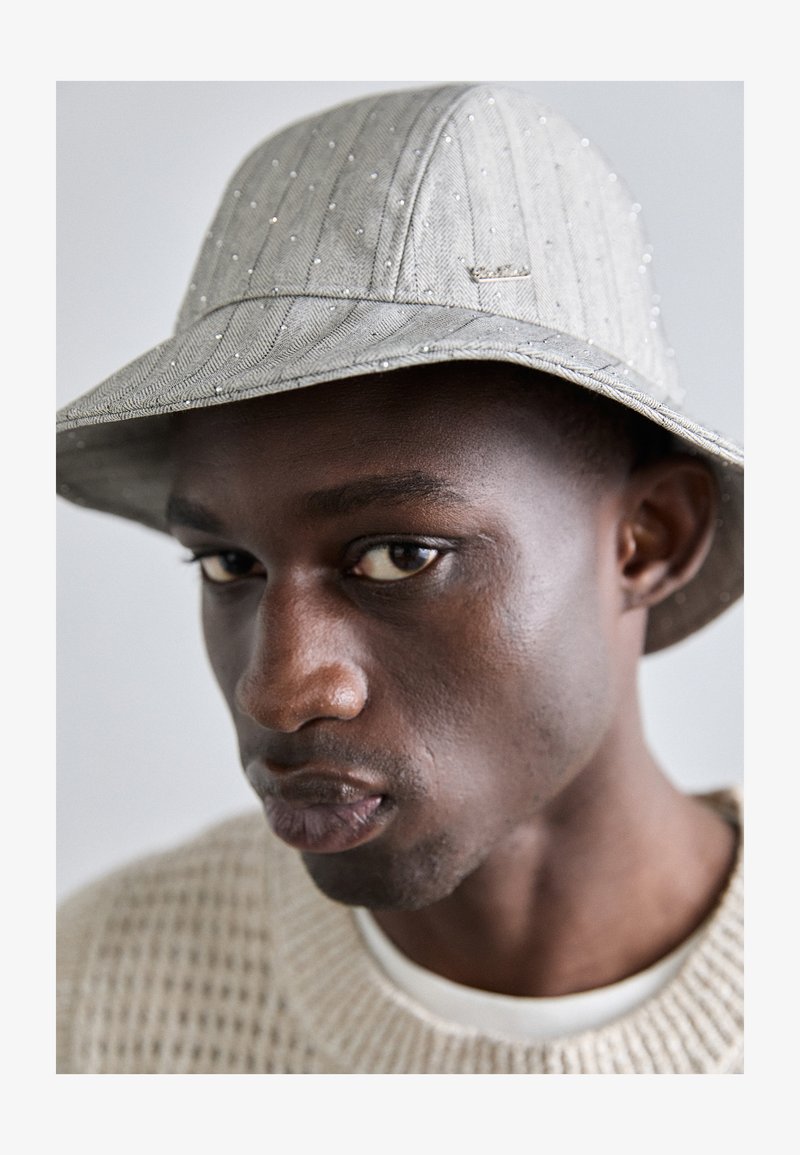 Hall gray bucket hat, mille peene triibud, kaunistatud väikeste läikivate aksessuaaridega. Serv on veidi allapoole keerdunud, koos tekstuuriga kootud sviitriga.