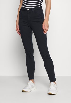 Woman wearing black skinny jeans, white sneakers, and a black and white striped sleeveless top standing against a plain white background.