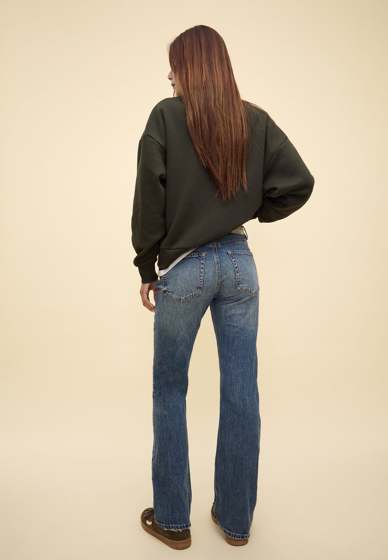 Donna con capelli castani lunghi che indossa un maglione verde scuro, jeans blu e scarpe marroni, in piedi con la schiena rivolta verso la macchina fotografica.