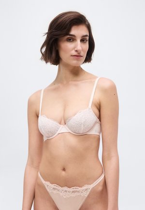 Kobieta o krótkich brązowych włosach, ubrana w bladoróżowy koronkowy biustonosz i pasujące koronkowe stringi na jasnym, jednolitym tle.