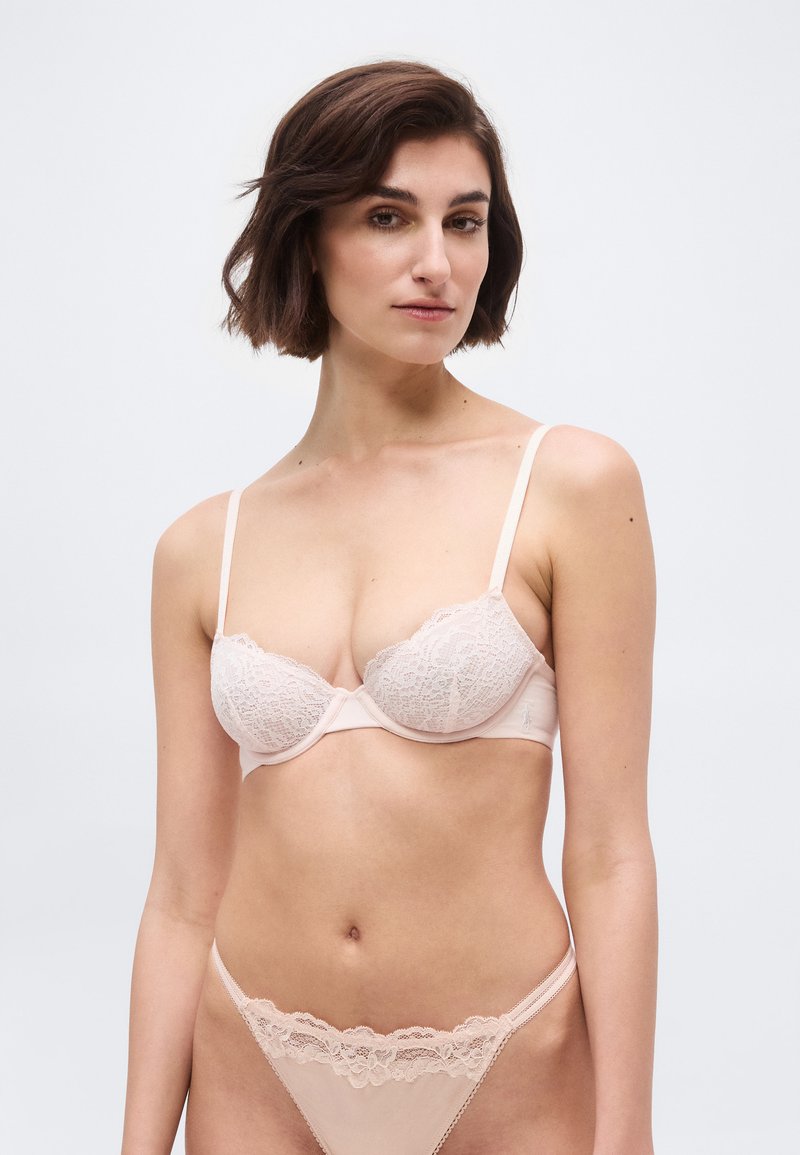 Kobieta o krótkich brązowych włosach, ubrana w bladoróżowy koronkowy biustonosz i pasujące koronkowe stringi na jasnym, jednolitym tle.