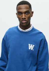 Sweatshirt bleu en coton, avec un col ras du cou et des poignets côtelés. Affiche un logo brodé blanc sur la poitrine gauche.