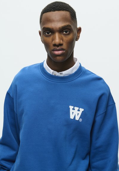 Sudadera azul de algodón, con cuello redondo y puños de canalé. Muestra un logo bordado en blanco en el lado izquierdo del pecho.