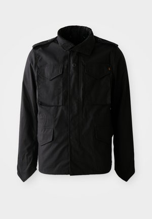 Chaqueta negra para hombre con cuatro bolsillos delanteros con tapa, con epaulettes abotonados y un cuello alto, exhibida sobre un fondo blanco.