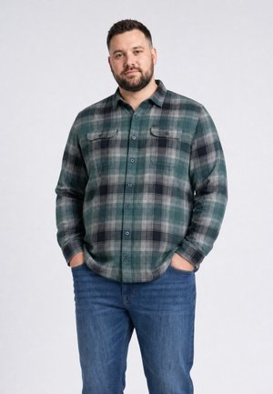 Mann mit Bart, der ein grün-grau kariertes Hemd und blaue Jeans trägt, steht mit den Händen in den Taschen vor einem schlichten Hintergrund.