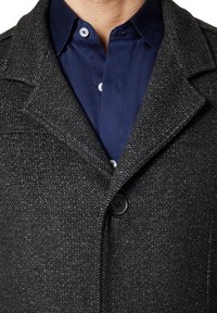 Szary wełniany płaszcz w wzór jodełki, z czarnym guzikiem i klapami, nałożony na niebieską koszulę z guzikami.