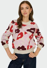 Pull en tricot présentant un motif floral rouge, bordeaux et rose sur un fond crème. Encolure et poignets côtelés. Coupe classique.