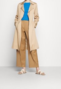 Trinchera beige con gran cuello, camiseta azul ajustada y pantalones de pierna ancha color tostado. Sandalias ligeras con múltiples tiras. Texturas de tela suaves.