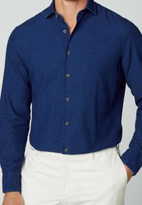 Hombre vistiendo una camisa azul oscuro ajustada abotonada, metida dentro de pantalones blancos, con una mano en el bolsillo, posando frente a un fondo liso.