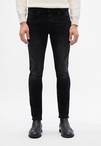 Jeans de denim negro con un corte slim, que cuentan con un diseño de cinco bolsillos, textura desgastada y detalles de costura en las costuras, combinados con botas negras.