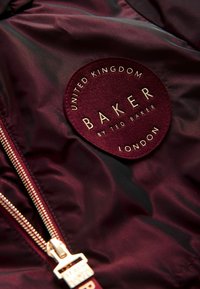 Veste en satin bordeaux avec un patch en feutre rond portant une inscription dorée : "BAKER par TED BAKER" et "ROYAUME-UNI LONDRES". Accent de fermeture éclair.