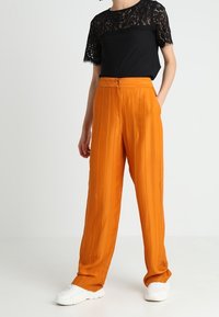Top de encaje negro de manga corta combinado con unos pantalones naranjas plisados de tiro alto. La modelo lleva zapatillas blancas. Fondo neutro.