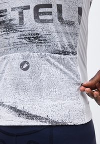 Egy személy fehér mintás kerékpáros mezben, kis skorpió logóval, zsebében lévő cipzárt igazít.