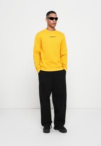 Yellow sweatshirt with a round neckline, paired with black cargo pants featuring side pockets. Model wearing black sunglasses and sneakers.