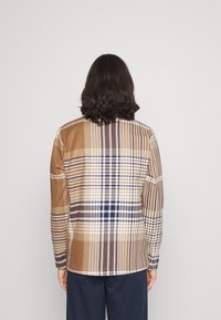 Camisa de manga larga de cuadros con una mezcla de colores marrón, beige y azul, con una textura tejida y patrones de cuadros en contraste en la parte trasera.