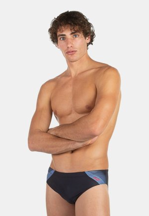 Giovane uomo con capelli castani ricci sta in piedi a braccia incrociate, indossando uno slip da bagno nero e blu su sfondo bianco semplice.