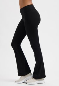 Schwarze, ausgestellte Leggings mit einem hochgeschnittenen Design, gefertigt aus einem glatten, dehnbaren Stoff. Kombiniert mit hellen Sportschuhen.