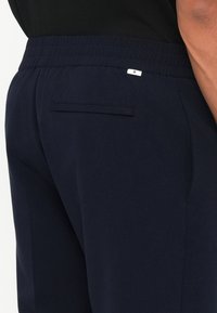 Calças azul marinho com cintura elástica, apresentando um bolso traseiro liso e uma pequena etiqueta da marca. Textura suave e forma ajustada.